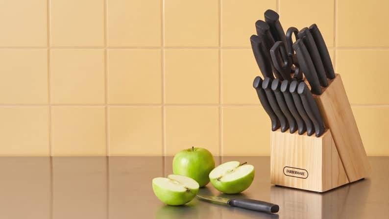 A wooden knife block with a set of black-handled knives next to green apples on a counter. Reads: From one seventy-six. Save on kitchen knives.